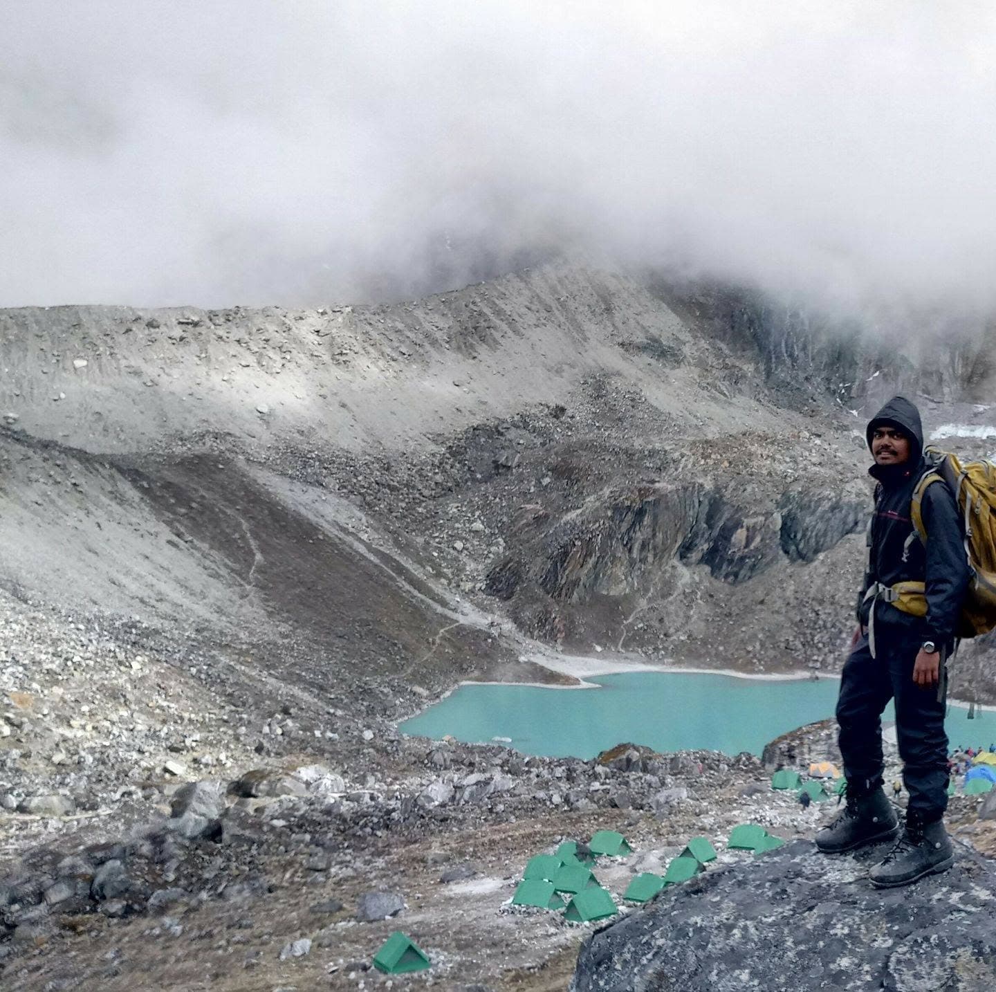 Kaushal Khodifad, Founder of CLOZOM, in full expedition kit with his hood up and a large pack, standing on a rock at the edge of a base camp. Below him, rows of green tents on a glacial moraine beside a turquoise meltwater lake, under flat cloud.